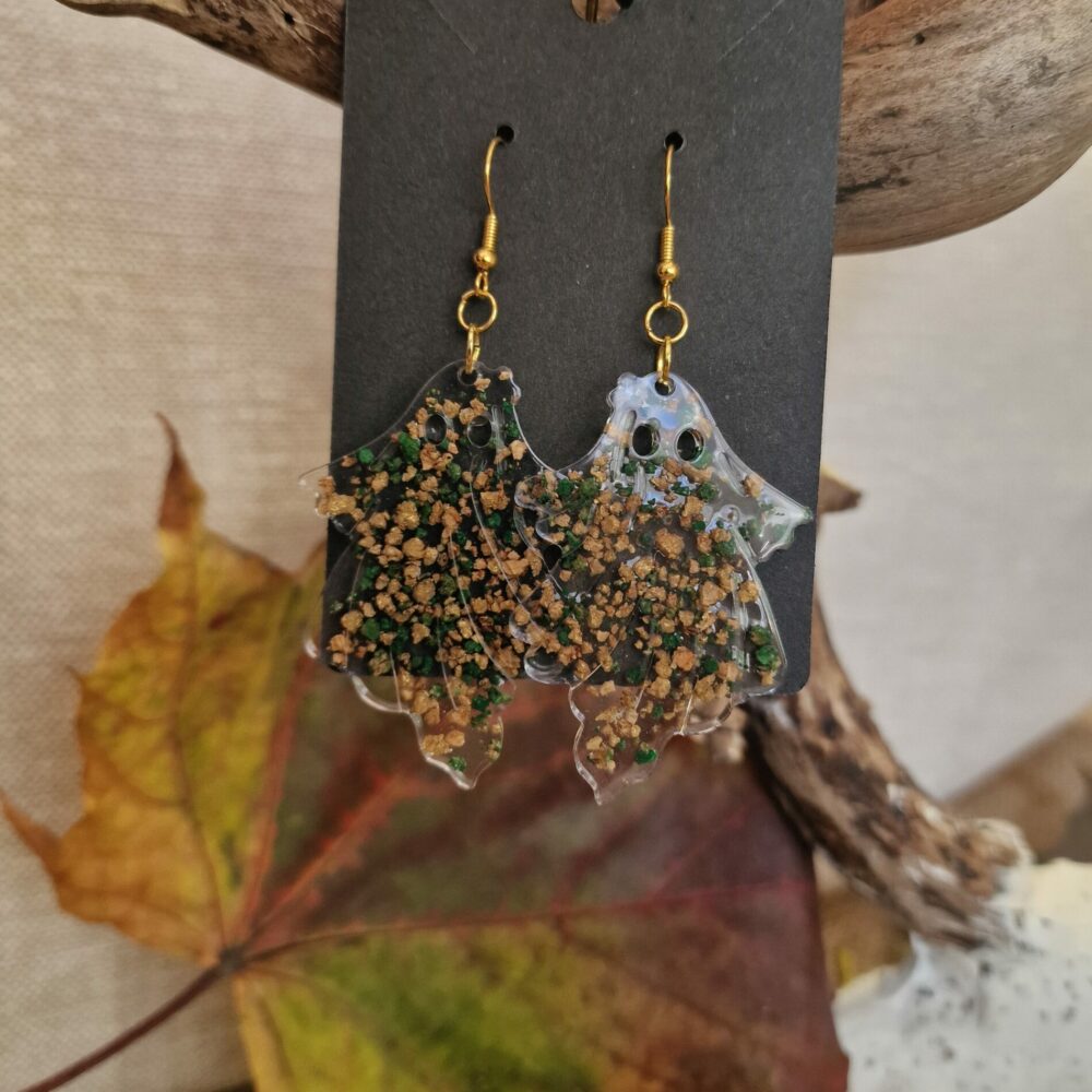 Boucles d’oreilles artisanales Cé’rénité en forme de petits fantômes, réalisées à la main en résine transparente avec inclusions de liège et éclats verts, attaches dorées.