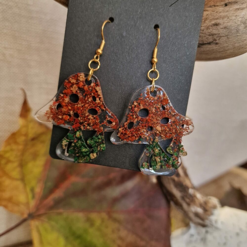Boucles d’oreilles artisanales Cé’rénité en forme de champignons, réalisées en résine transparente avec inclusions de liège et éclats verts et rouges, attaches dorées.