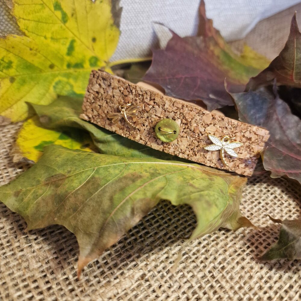 Pince à cheveux artisanale en liège décorée d’un bouton vert et de deux libellules dorées, présentée sur fond de feuilles d’automne.
