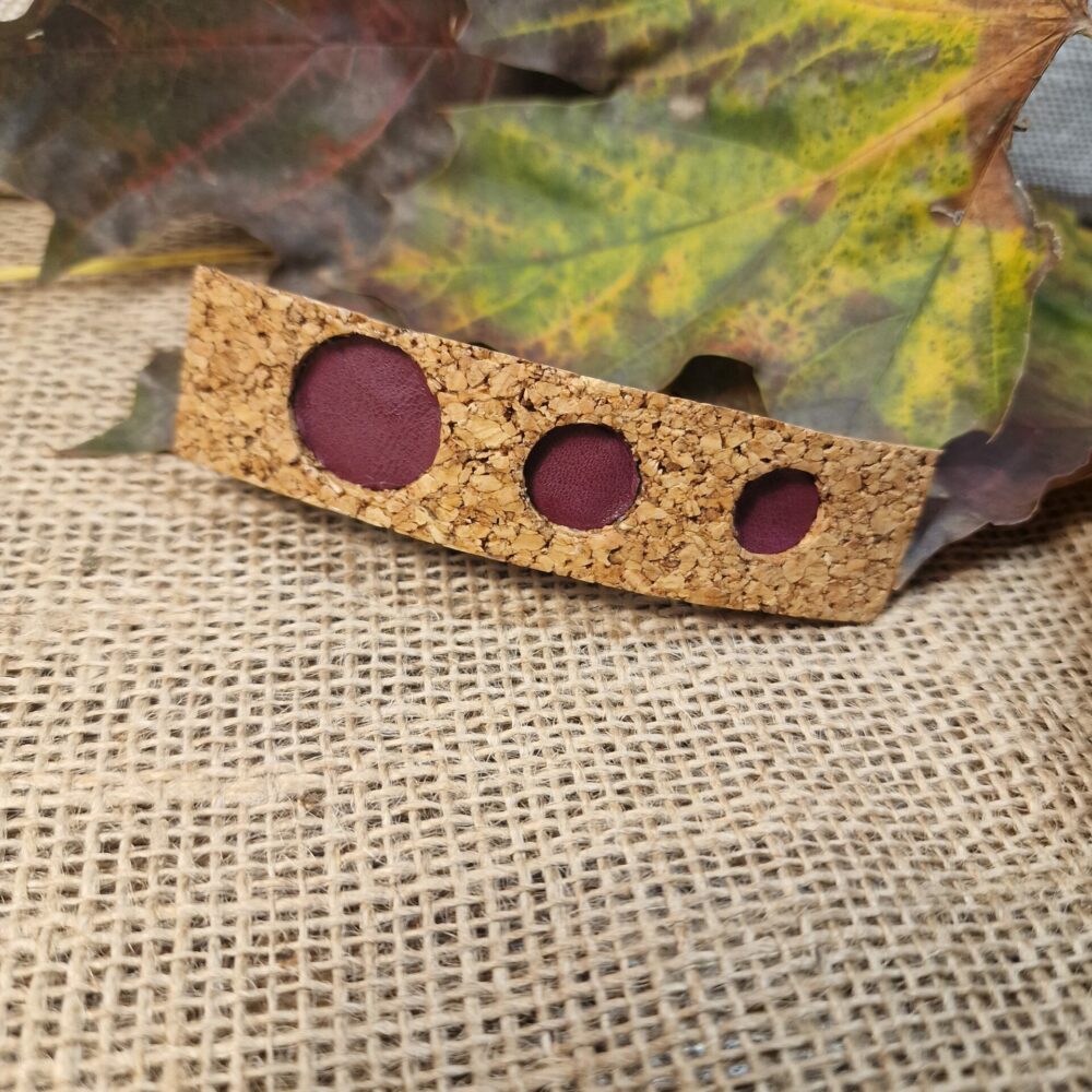Pince à cheveux artisanale en liège décorée de trois pastilles en cuir bordeaux mauve, présentée sur fond de feuilles d’automne.