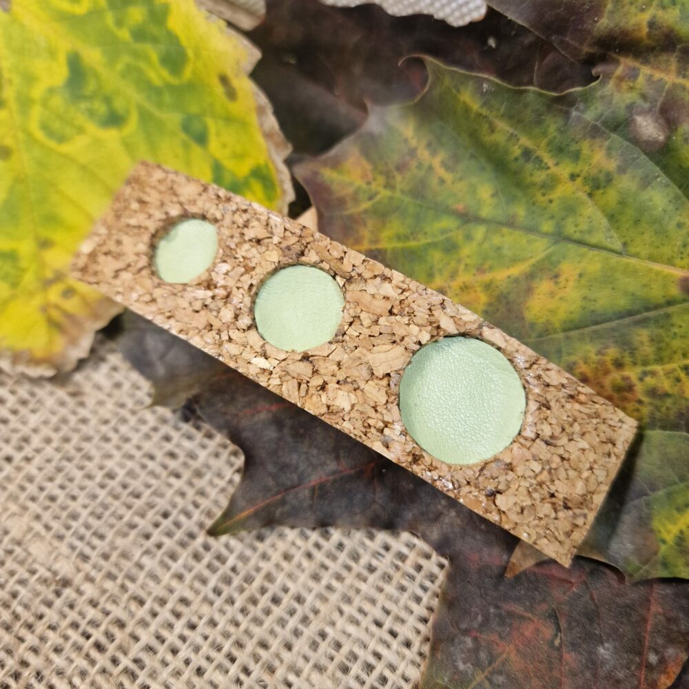 Pince à cheveux artisanale en liège décorée de trois ronds en cuir vert clair, présentée sur fond de feuilles d’automne.